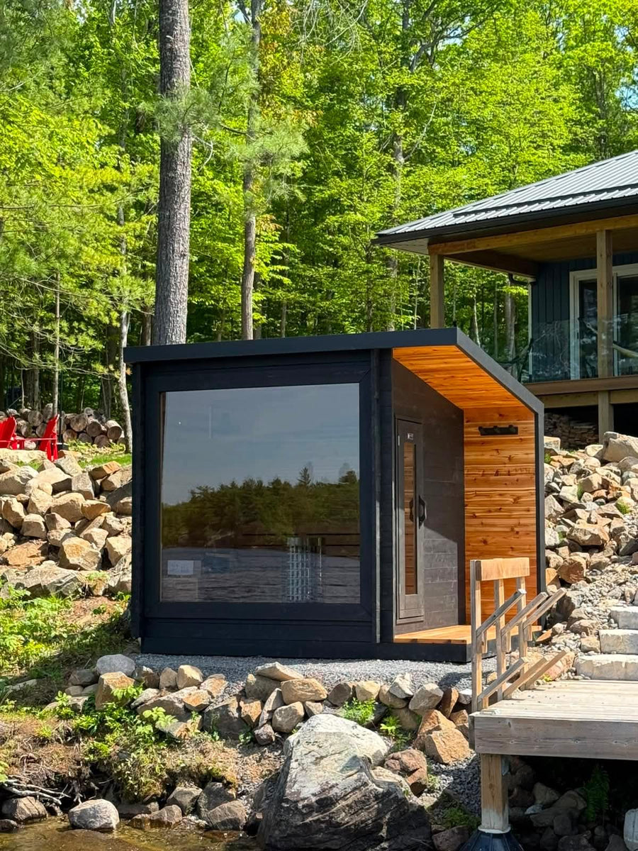 A black cedar outdoor sauna with a full glass wall and covered porch on a rocky lake shore